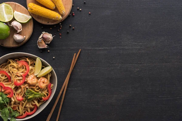 Вид сверху на вкусную пряную тайскую лапшу рядом с палочками для еды и свежие ингредиенты на деревянной серой поверхности с копировальным пространством — стоковое фото