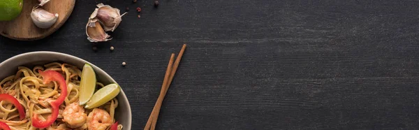 Vista dall'alto di gustose tagliatelle thai piccanti con aglio e gamberetti vicino a bacchette su superficie grigio legno, colpo panoramico — Foto stock