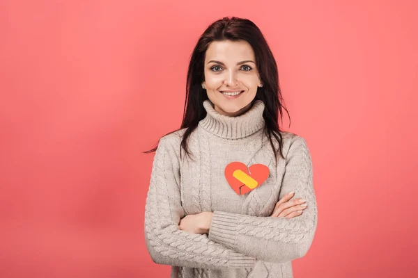 Mujer alegre con tirita amarilla sobre papel en forma de corazón cortado de pie con los brazos cruzados en rosa - foto de stock