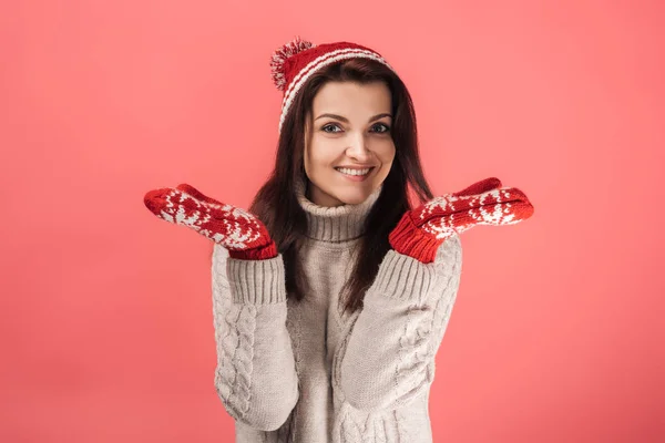 Mujer feliz en guantes rojos y sombrero de punto mostrando gesto de encogimiento de hombros en rosa - foto de stock