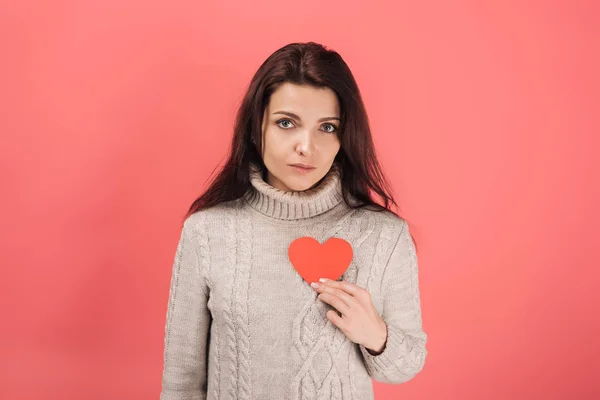 Upset woman in sweater holding heart-shaped paper cut on pink — Stock Photo