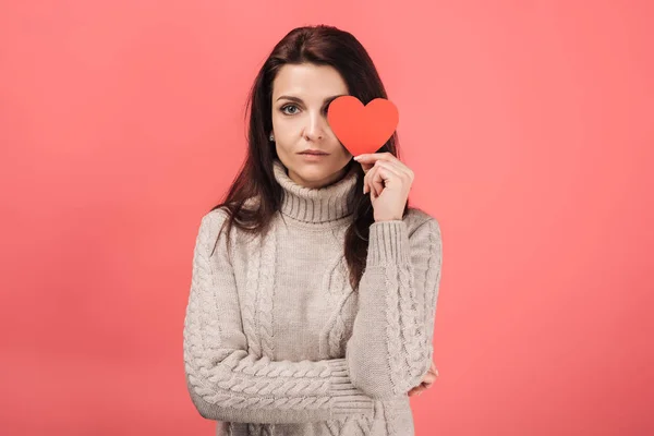 Mujer en suéter cálido que cubre el ojo con papel en forma de corazón cortado en rosa - foto de stock