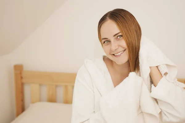 Lovely female with wet hair, dries head with towel, being pleased after taking bath, dressed in white bathrobe