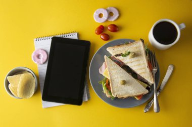 Atıştırmalıklar, fast food konsepti. İş yerinde yemek yiyor. Taze sossandviç, sebze, kahve, patates cipsi, tatlı kurabiye. Tablet ekranı. Sarı arka plan.
