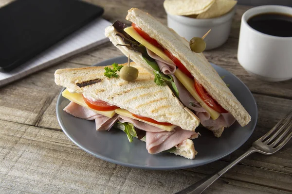 Comer insalubre en el trabajo. Triángulo, sándwich club con jamón y verduras. Tableta, pantalla de gadget y cuaderno. Una taza de café. Comida rápida . — Foto de Stock