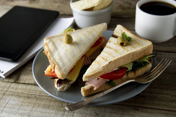 Comer insalubre en el trabajo. Triángulo, sándwich club con jamón y verduras. Tableta, pantalla de gadget y cuaderno. Una taza de café. Comida rápida . — Foto de Stock