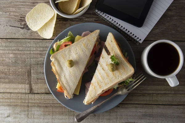 Comer insalubre en el trabajo. Triángulo, sándwich club con jamón y verduras. Tableta, pantalla de gadget y cuaderno. Una taza de café. Comida rápida . — Foto de Stock