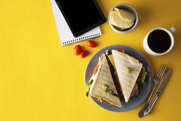 Snacks, concepto de comida rápida. Comer en el trabajo. Sandwich de club fresco, verduras, café, papas fritas, galletas dulces. Pantalla de tableta. Fondo amarillo . — Foto de Stock