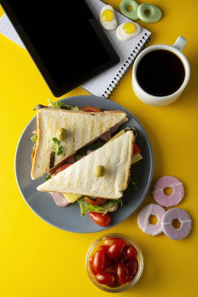 Snacks, concepto de comida rápida. Comer en el trabajo. Sandwich de club fresco, verduras, café, papas fritas, galletas dulces. Pantalla de tableta. Fondo amarillo . — Foto de Stock