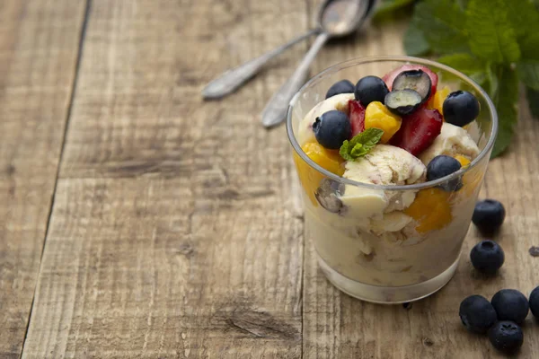 Vanilla ice cream with fruits - blueberries, strawberries, mango and mint, summer refreshing dessert. Wooden background. Glass with vanilla icecream.