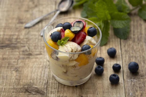 Fruit salad with ice cream - blueberries, strawberries, mango and mint, summer refreshing dessert. Wooden background. Glass with vanilla icecream.
