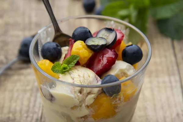 Fruit salad with ice cream - blueberries, strawberries, mango and mint, summer refreshing dessert. Wooden background. Glass with vanilla icecream.