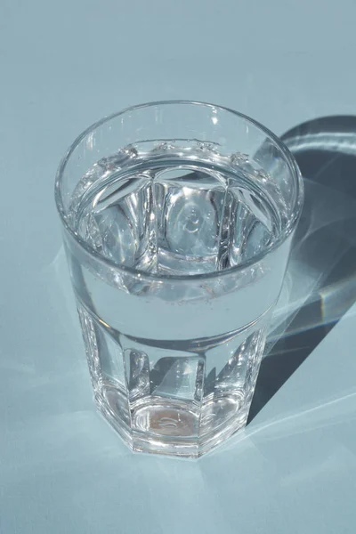 Um copo de água pura sob a luz solar com profundas sombras elegantes no espaço de cópia de fundo azul — Fotografia de Stock