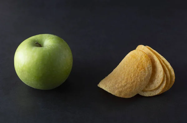 Food choise concept. Potatoe chips or green apple for snack. Top view, black background.