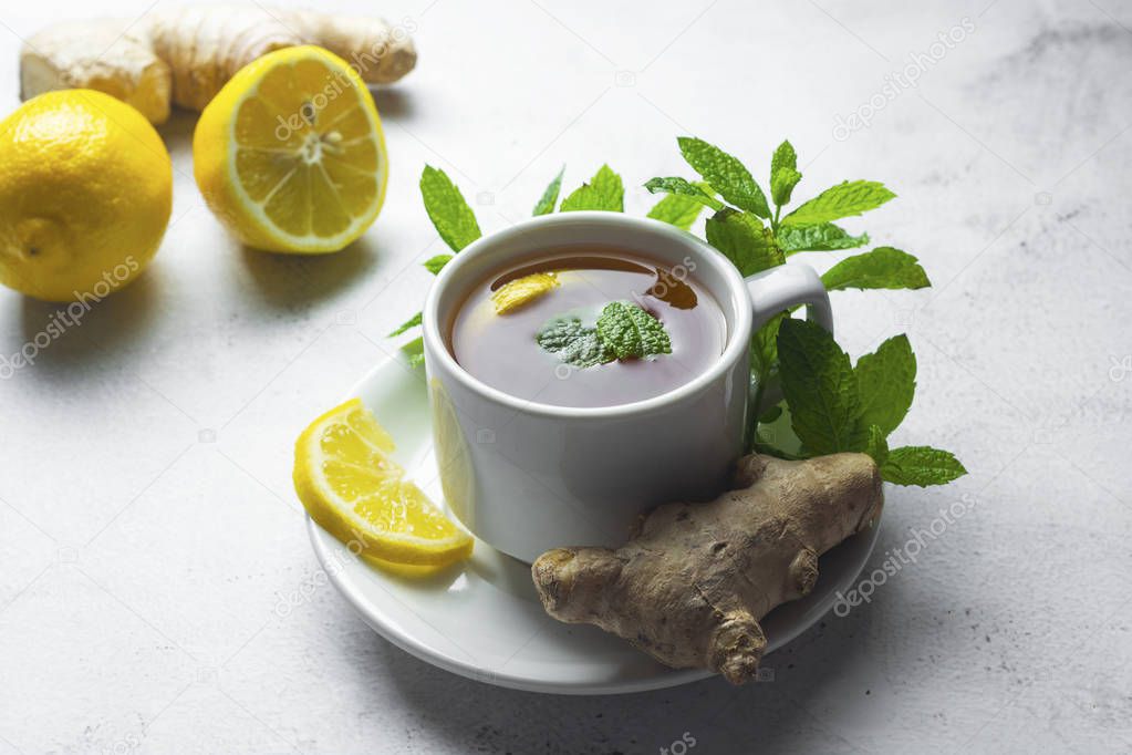 Glass cup of ginger tea with lemons and mint leaves on light background. Ginger tea, drink ingredients, cold and autumn time.