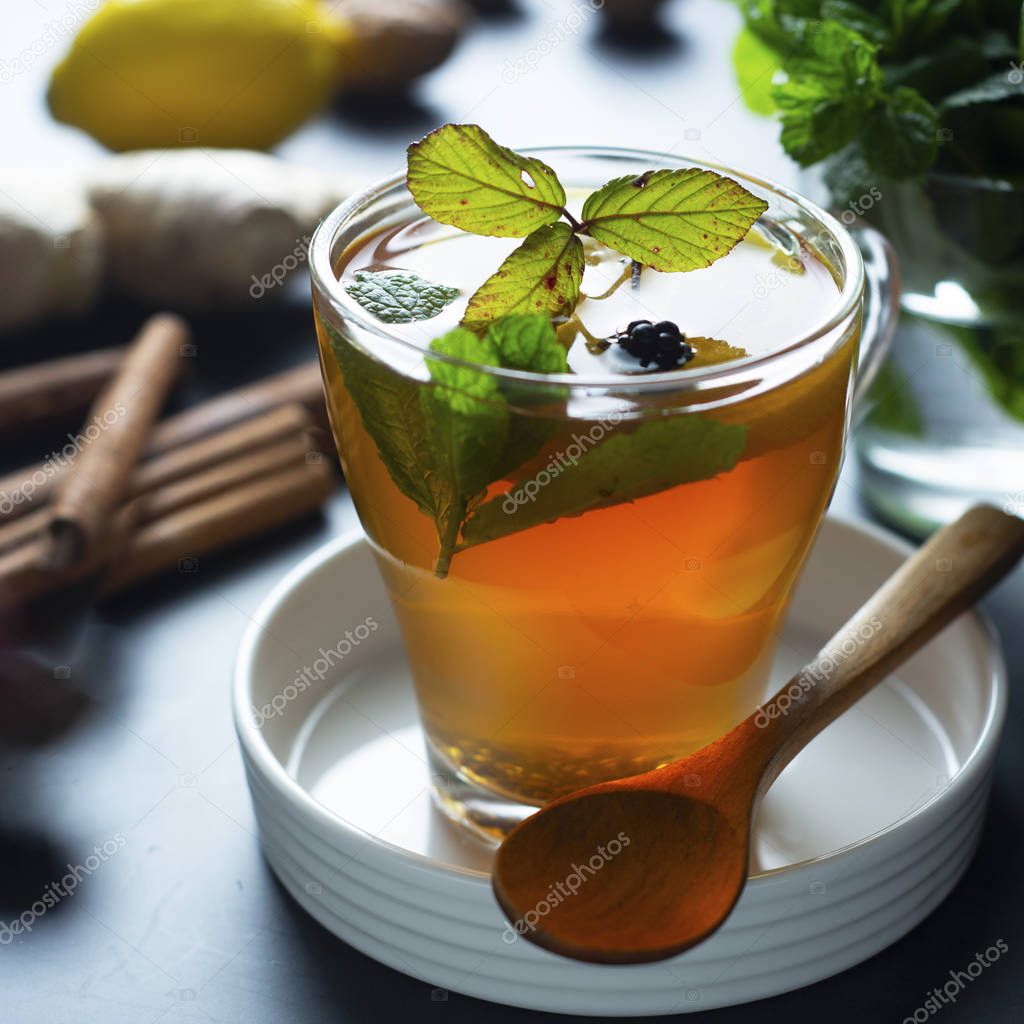 Glass cup of ginger tea with lemons and mint leaves on dark background. Square image, cold and autumn time.
