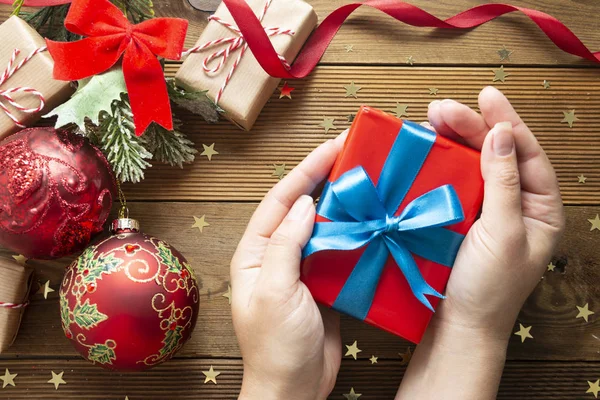 Kvinnliga händer håller julklapp, röd presentask. Förpackning insvept presentförpackning på trä bakgrund. Julvintern dekorationer, röda grannlåt, gran grenar, röd rosett. — Stockfoto