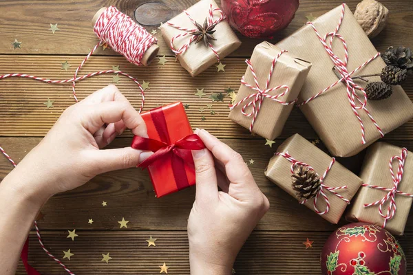 Kvinnliga händer håller, inslagning jul presentförpackning. En grupp presentlådor wraped i hantverk papper, röda grannlåt, glitter över träbord. Chritsmas platt låg bakgrund. — Stockfoto