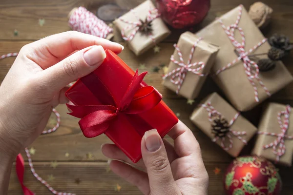 Női kéz a kézben, karácsonyi ajándékdoboz becsomagolva. Ajándékdobozok csoportja, kézműves papírba csomagolva, piros dísztárgyak, csillámpor fából készült asztalokon. Chritsmas lapos feküdt háttér. — Stock Fotó