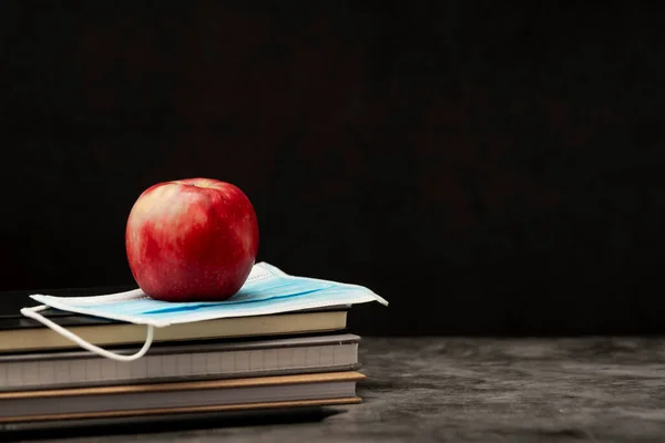 Stack of note books, red apple and face protective medical mask. Black board with copy space.