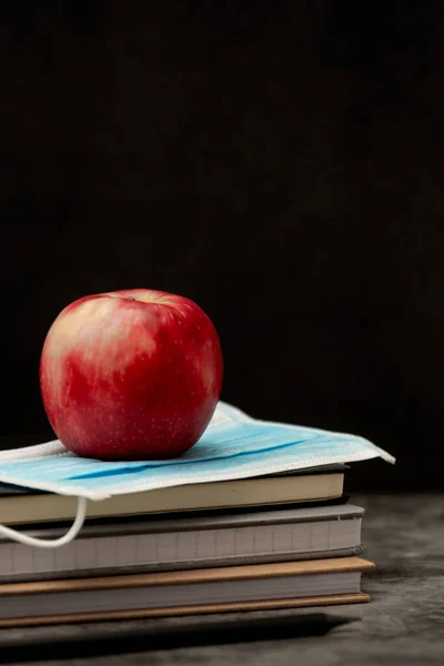 Stack of note books, red apple and face protective medical mask. Black board with copy space.