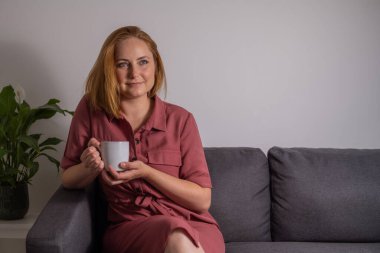 Orta yaşlı sarışın kadın, evinde kanepede kahve içiyor.