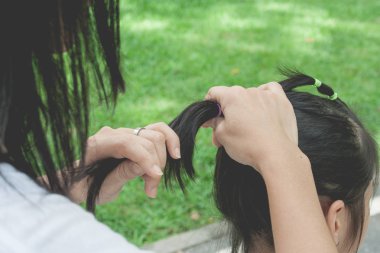 Woman tying black hair of little cute child in ponytail style with elastic band at public park. clipart