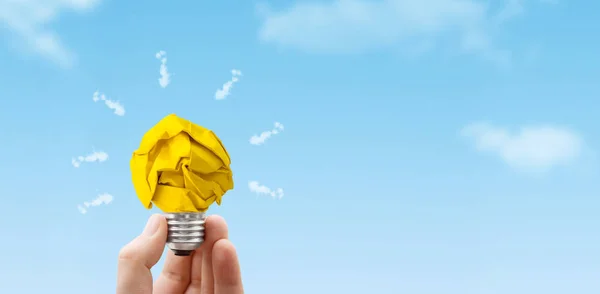 Business Idea Concept : Hand holding light bulb made from yellow crumpled paper ball with blue sky in background.