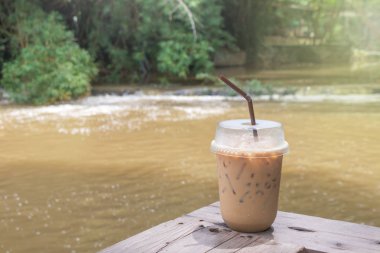 Su akıntısı ve arka planda yeşil ağaçlar olan ahşap masanın üzerinde plastik bardakta buzlu kahve ve saman..