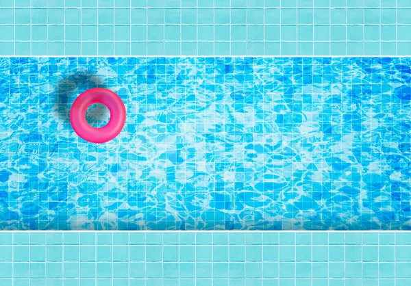 Pink inflatable floating ring floating on blue water in swimming pool surrounded with tiles mosaic floor from top view.