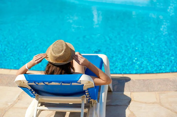 Woman in a straw hat relaxing on a deck-chair near a luxurious summer pool, concept time to travel — Stock Photo, Image