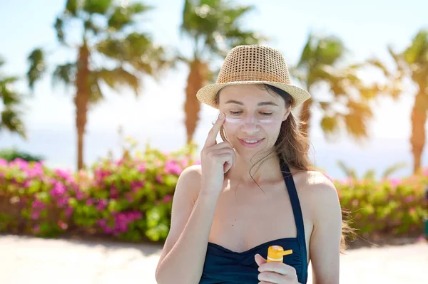 Portrait of a beautiful young woman, smiling in a swimsuit smeared face with sun protection cream in hat on sea background. Concept: sea sun and travel vacation freedom, enjoy sunburn heat hot — Stock Photo, Image