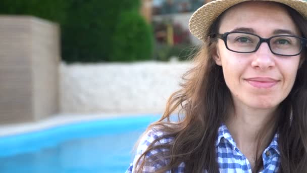 Das Profil einer jungen, hübschen Frau mit Sonnenbrille, die am Pool sitzt und sich an einem Sommertag in einem stylischen Badeanzug sonnt, im Hintergrund leuchtet das Poolwasser — Stockvideo