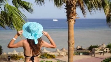Güzel kadın sahilde dinlenme ve denize bakarak. Kız şapkasını düzeltiyor ve rahat yerleşir