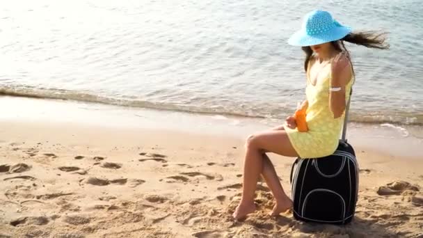 Mooie vrouw in een hoed met een pot van zonnebrand crème in haar handen zit op een koffer tegen de zee en haar schouder uitstrijkjes tijdens zomervakantie reizen. Mooie gelukkige vrouw met zonne-olie crème in — Stockvideo