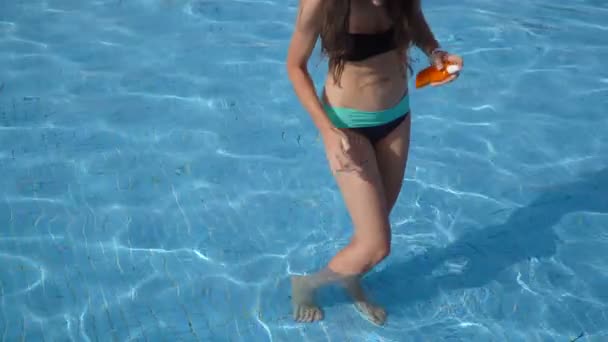 美丽的女人在比基尼与一罐防晒霜在她的手在游泳池的水在夏季旅游假期。美丽的快乐的妇女与太阳油奶油在塑料喷雾瓶在海滩上 — 图库视频影像