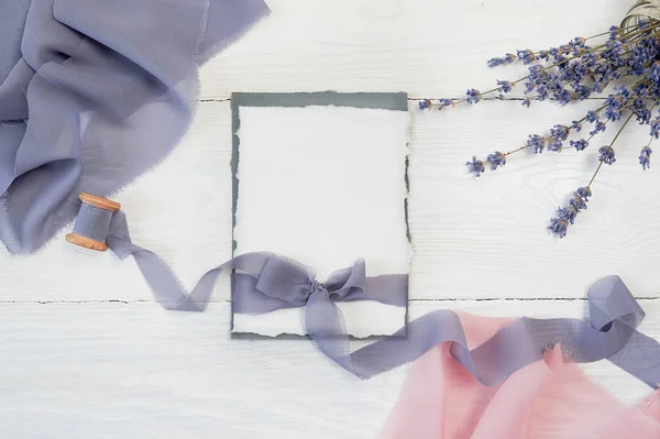 Weiße leere Karte Schleife auf einem Hintergrund aus rosa und blauen Stoff mit Lavendelblüten auf weißem Hintergrund. Attrappe mit Umschlag und Blanko-Karte. flach lag. Ansicht von oben — Stockfoto