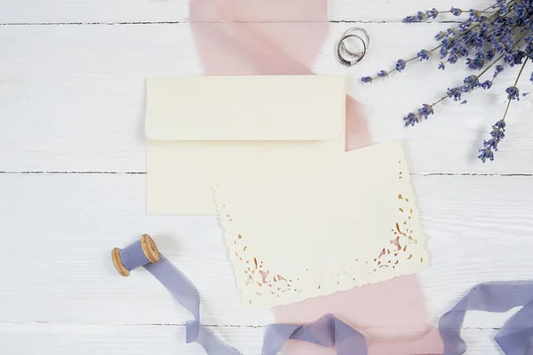 White blank card, envelope and ribbon with two wedding rings on a background of pink fabric with lavender flowers on a white background. Mockup with envelope and blank card. Flat lay. Top view