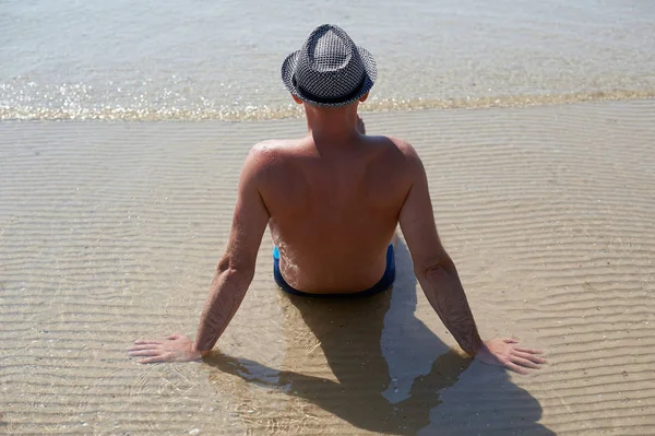 Portret styl życia całkiem młody człowiek opalone w kapelusz lato. Cieszyć się życiem i siedząc na plaży, czas na podróż. Z widokiem na morze — Zdjęcie stockowe