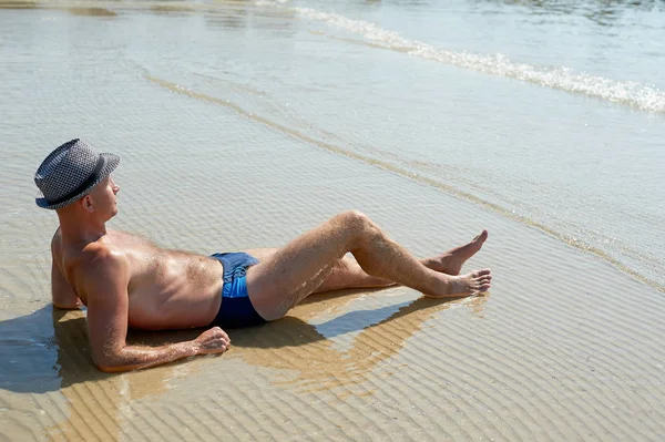Sommer-Lifestyle-Porträt eines hübschen jungen, sonnengebräunten Mannes mit Hut. Das Leben genießen und am Strand sitzen, Zeit zum Reisen. Blick auf das Meer — Stockfoto