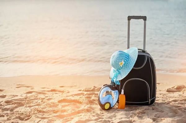 Suitcase and hat, sunscreen with a mask. The tropical sea, beach in the background. The concept of summer recreation travel and cruise traffic