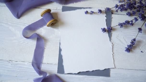 Carte blanche vierge, enveloppe et ruban sur fond de tissu rose et bleu avec des fleurs de lavande sur fond blanc. Maquette avec enveloppe et carte blanche. Pose plate. Vue du dessus — Video