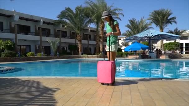 Молодая женщина с розовым футляром возле бассейна в отеле. Концепция туристического отпуска — стоковое видео