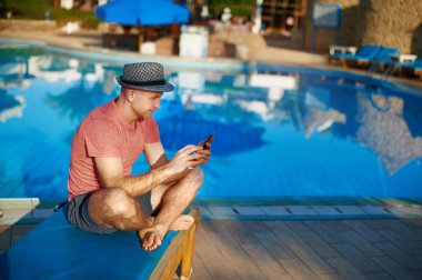 Genç adam dinlenme üzerinde güneş şezlong tarafından yüzme havuzu ve türü sms telefon, iş başarılı bir çevirmen