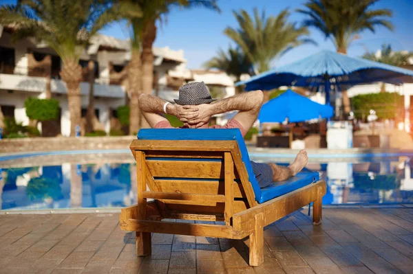 Hombre joven en sombrero se encuentra en más alto cerca de la piscina para relajarse concepto de vacaciones de verano — Foto de Stock