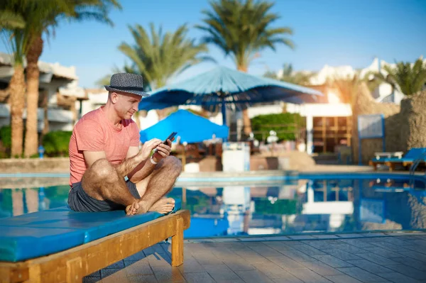 Jeune homme se reposant sur des chaises longues près de la piscine et le type sms sur le téléphone, freelancer travaille à distance du bureau — Photo