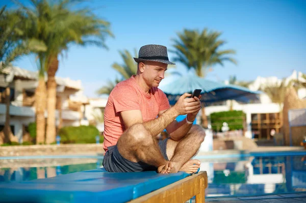 Junger Mann ruht sich auf Liegestühlen am Swimmingpool aus und tippt SMS am Telefon, freiberuflicher Job — Stockfoto