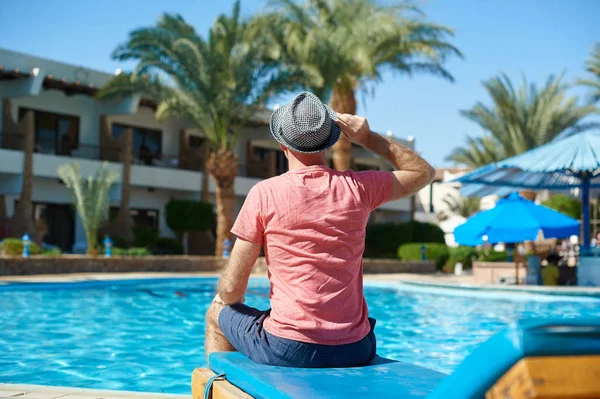 Jeune homme en chapeau assis sur des chaises longues près de la piscine hôtel — Photo