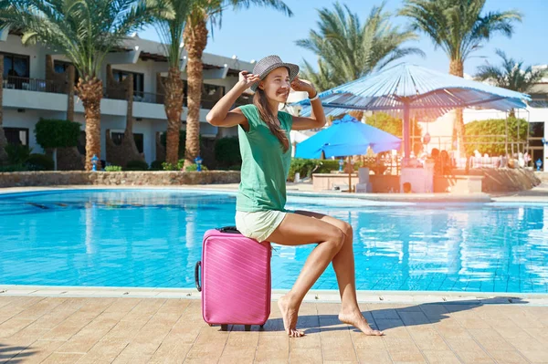 Женщина сидит на розовом чемодане рядом с бассейном отеля. Путешествия, летний отдых и концепция отпуска — стоковое фото