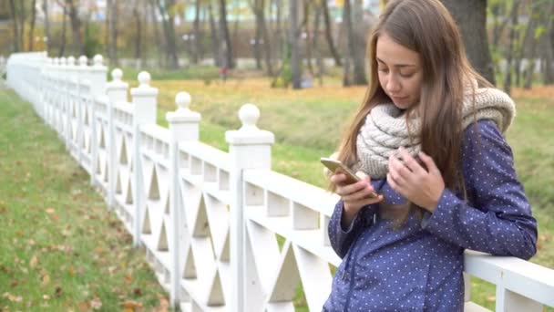 Кавказький жінка з knitten шарф, використовуючи смарт-телефону, набравши щось під час прогулянки у парку восени. відео 4 к — стокове відео